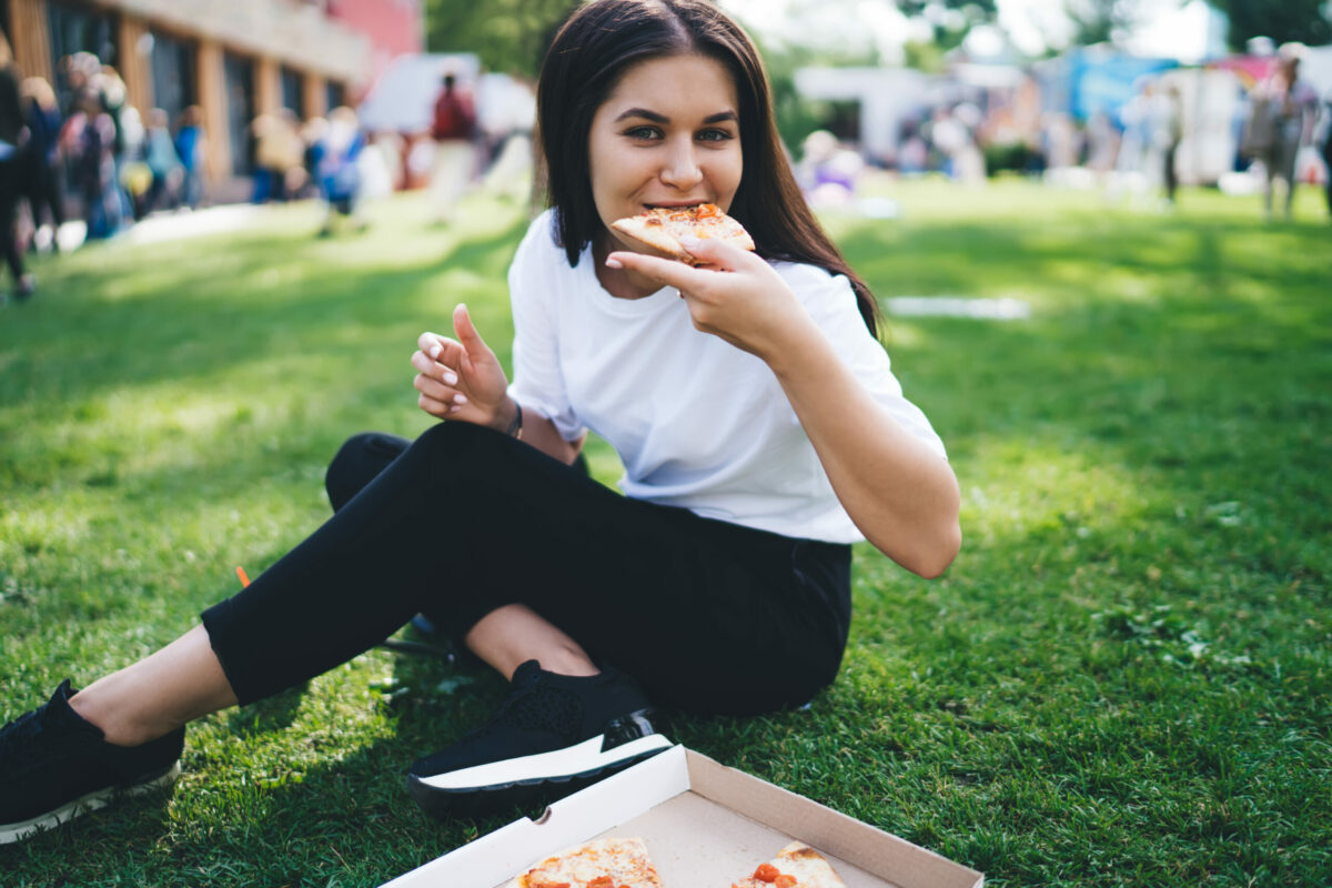 Hungry caucasian female in casual wear eating delicious italian pizza on weekend in park in city, beautiful hipster girl 20s satisfied with tasty fast food snack on lunch chilling during celebration