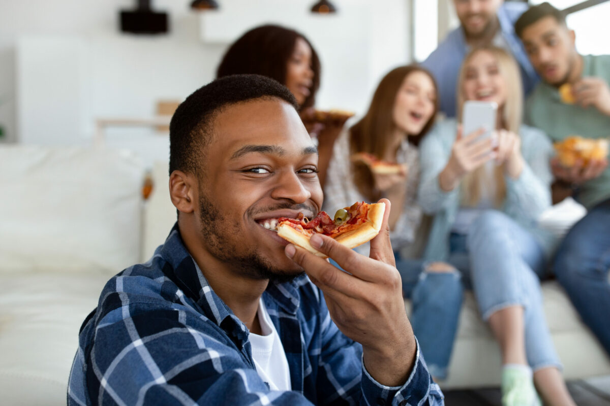 Attractive black guy eating yummy pizza, his diverse friends with cellphone having fun on background. Millennial students having social gathering, talking and laughing at home. People and lifestyles