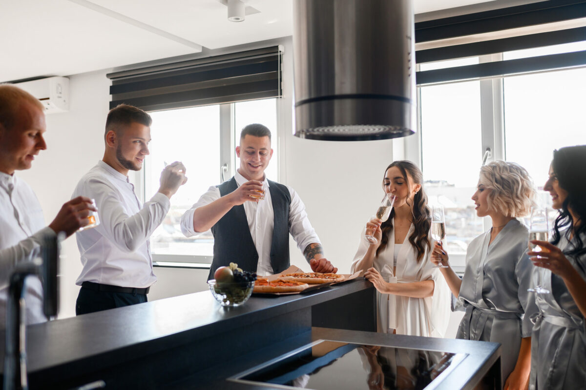 Front view of bride in a silk robe and groom in black suit celebrating wedding morning with bridesmaids and groomsman in spacious apartment in kitchen. Brides drinking with friends.